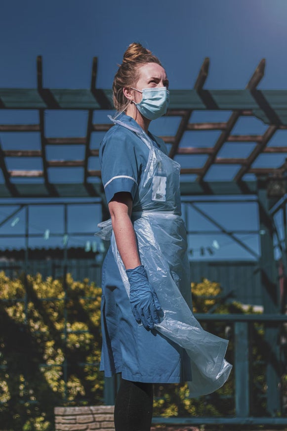 A woman wearing a blue medical face mask, scrubs, protective apron and gloves stands outside on a sunny day.